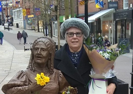 Dr Maureen Wright standing beside the statute of Elizabeth in Congleton. She is holding flowers.